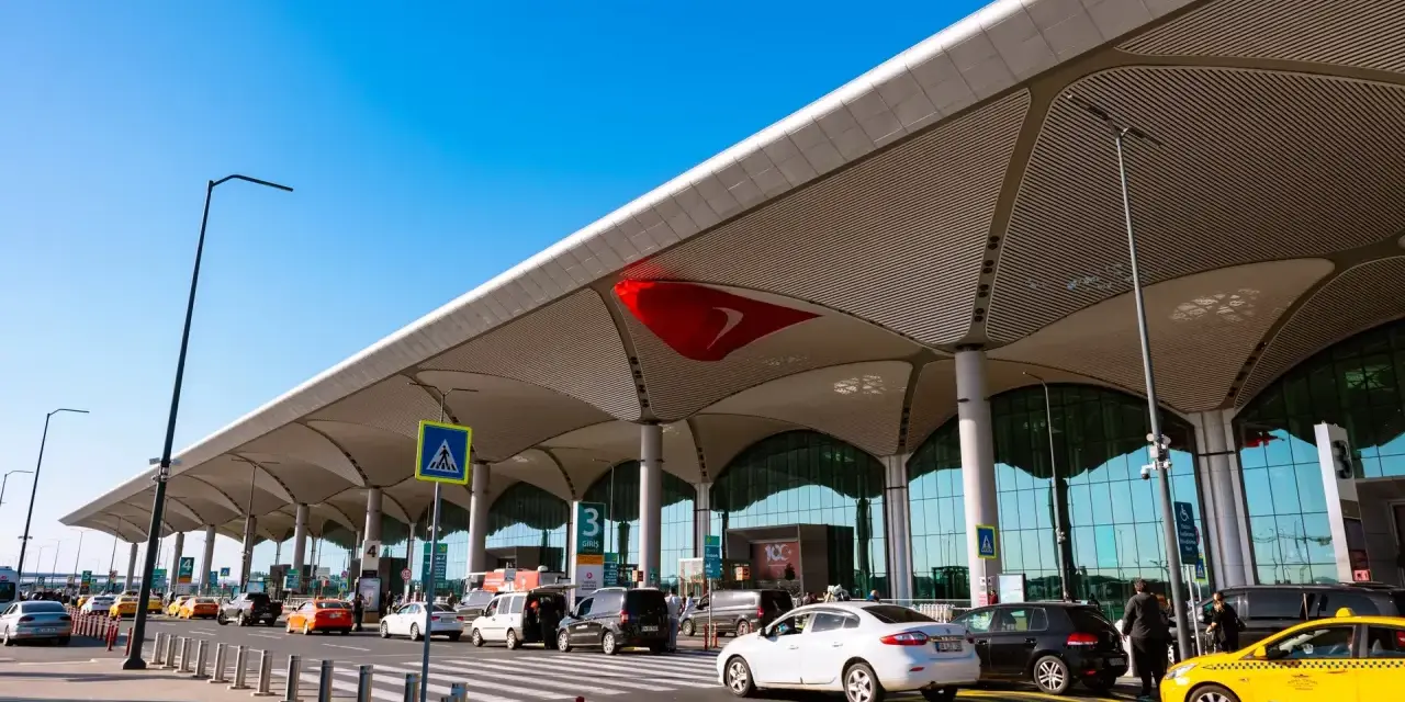 Nowoczesny budynek lotniska Bodrum z charakterystycznym zadaszeniem. Przed terminalem ruch samochodowy, w tym żółte taksówki.