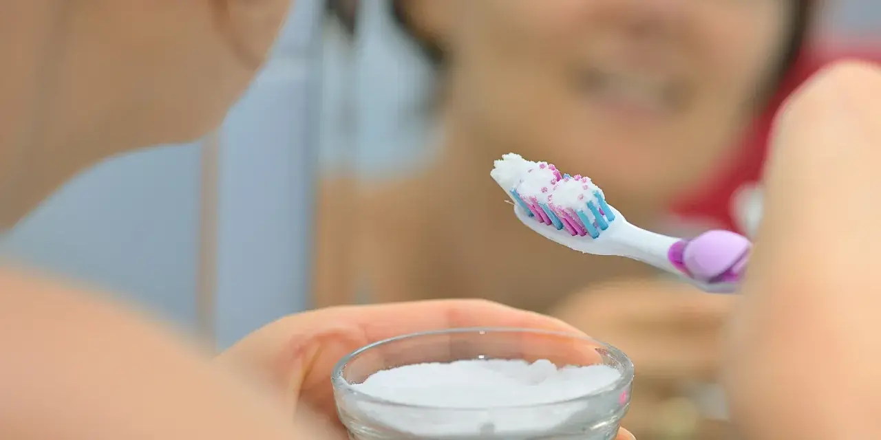 Ręka trzymająca szczoteczkę do zębów z białym proszkiem, obok miseczka z sodą oczyszczoną. Domowa pasta do zębów gotowa.