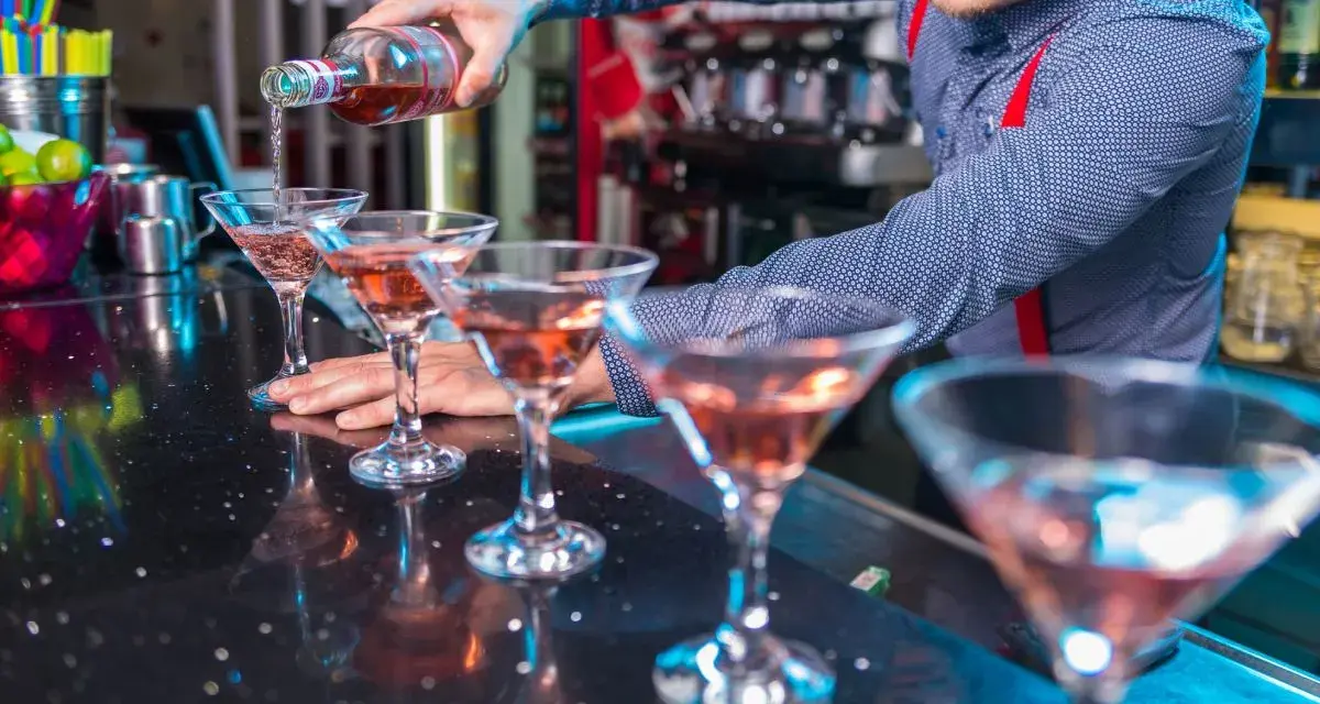 Barman nalewa różowy drink do kieliszków koktajlowych na czarnym blacie.
