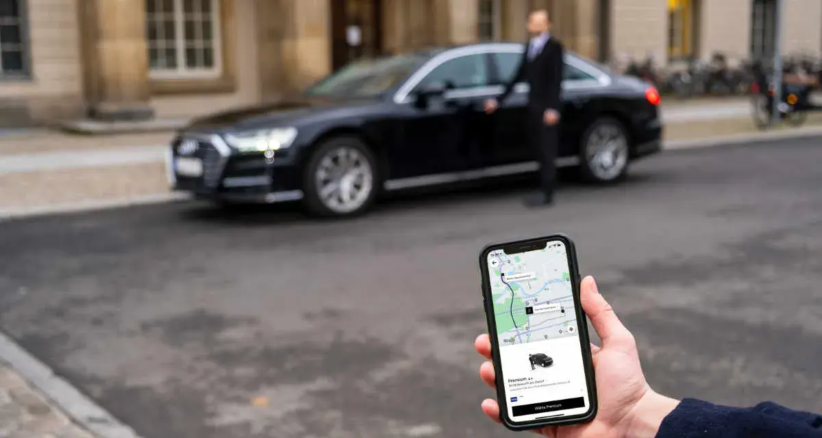 Hand hält Smartphone mit Uber-App, die eine Fahrt mit uber premium cars anzeigt. Ein Fahrer steht neben einem schwarzen Audi.