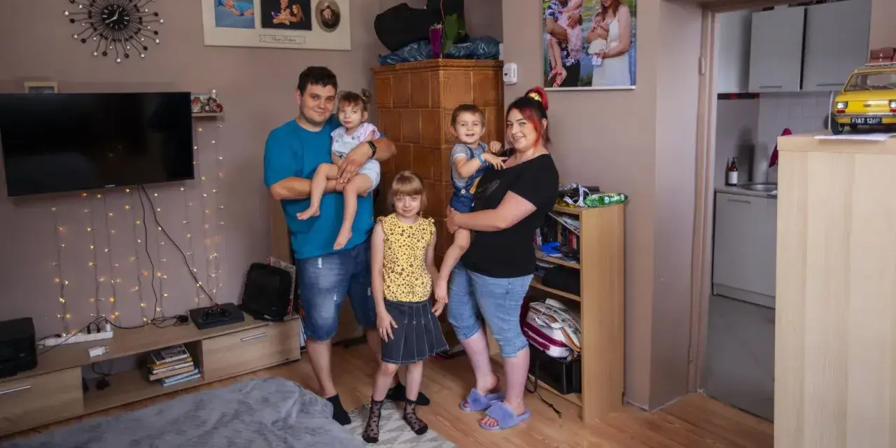 Rodzina z trójką dzieci pozuje do zdjęcia w salonie. W tle widać zdjęcia, telewizor i ozdobne lampki. Ich życie na kredyt nie przeszkadza im w cieszeniu się chwilą.