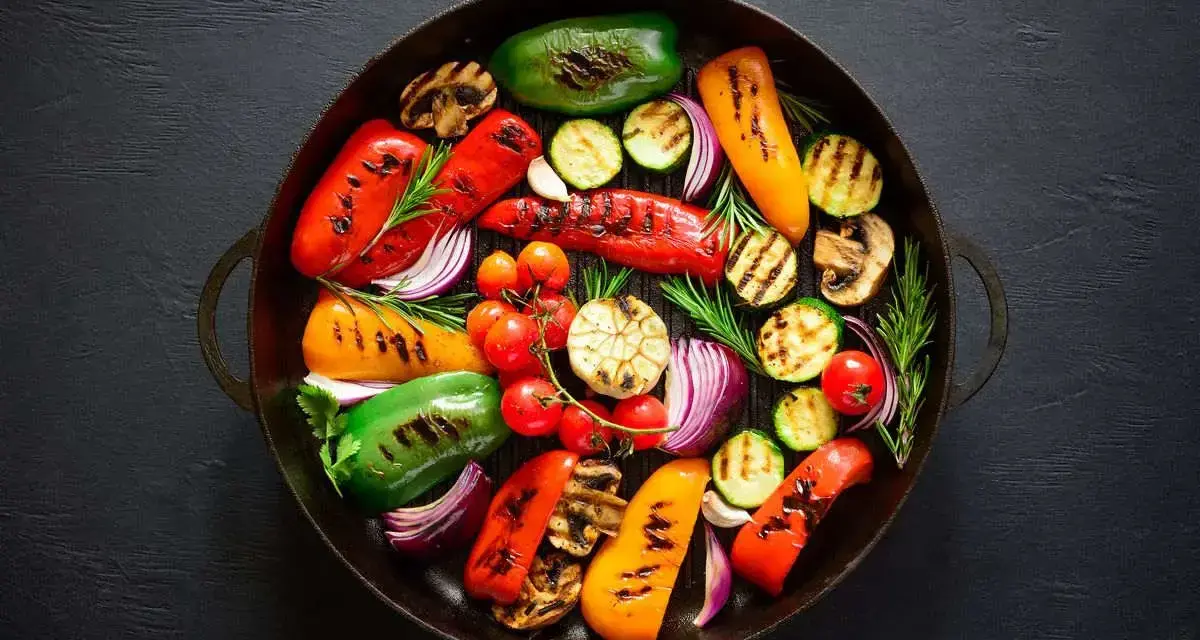 Un assortimento colorato di verdure grigliate, tra cui peperoni, zucchine, pomodorini, cipolle rosse e funghi, disposte in una padella scura.