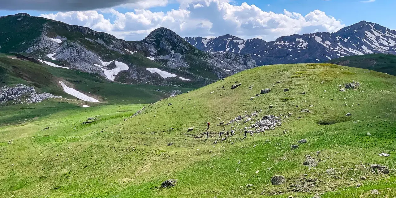 Grupa turystów wędruje po zielonych zboczach gór Macedonii, gdzie w oddali widać ośnieżone szczyty.
