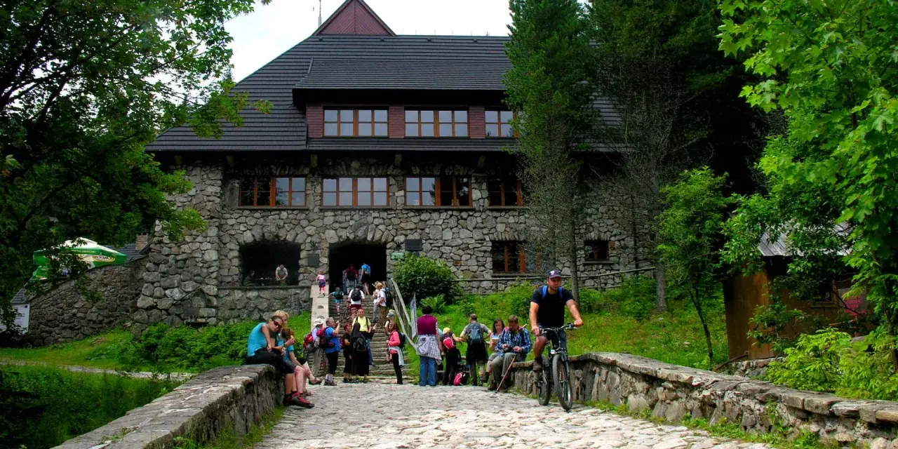 Kamienny budynek schroniska w Dolinie Chochołowskiej z ludźmi na schodach i rowerzystą na kamiennej drodze.