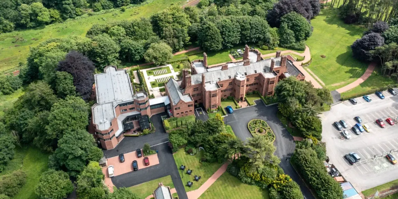 Aerial view of the abbey guest house, a grand brick building surrounded by lush green trees and manicured lawns. Cars are parked in a lot.