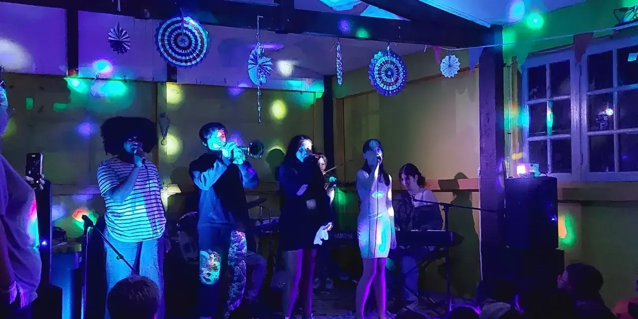 Scène de concert animée avec des lumières colorées. Un groupe joue, avec des chanteurs, un trompettiste et un claviériste. Le public, dont des enfants, filme le spectacle. Ces vacances musicales sans frontières sont une fête pour les sens.