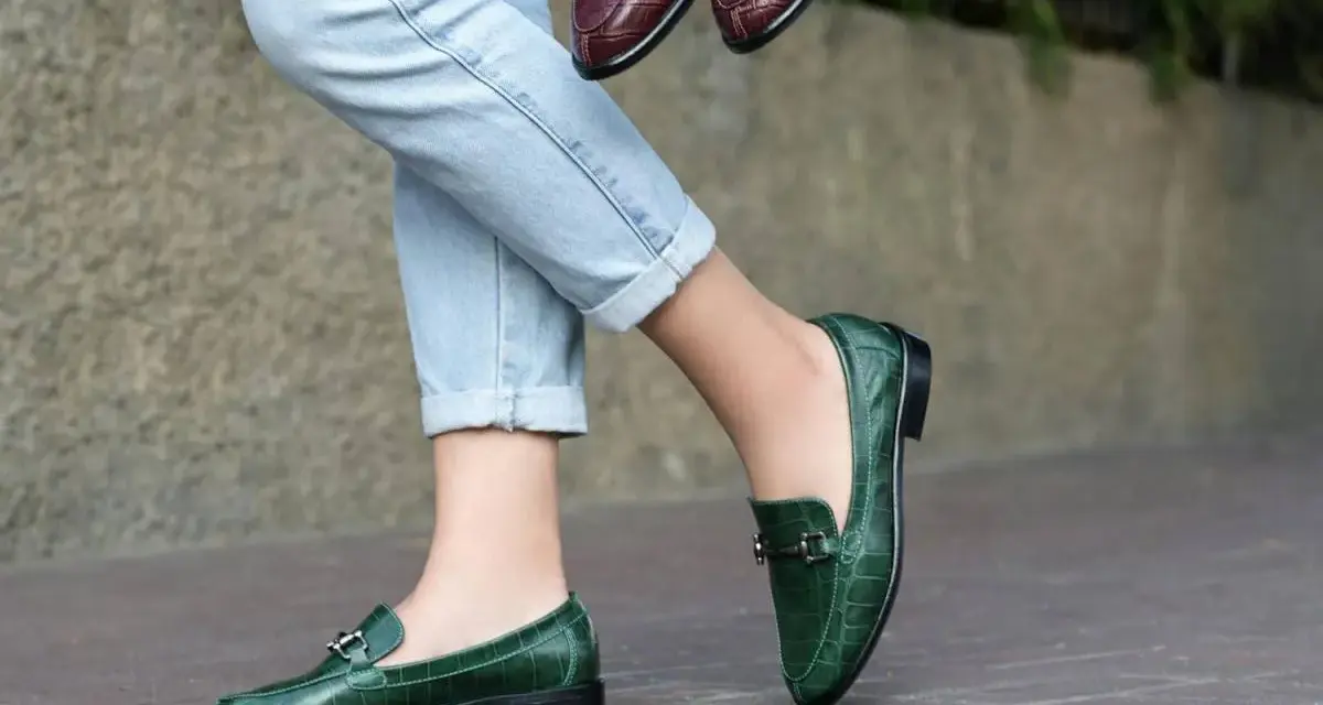 Un mocassino verde con abito elegante, perfetto per un look sofisticato. Un altro paio marrone è tenuto in mano.