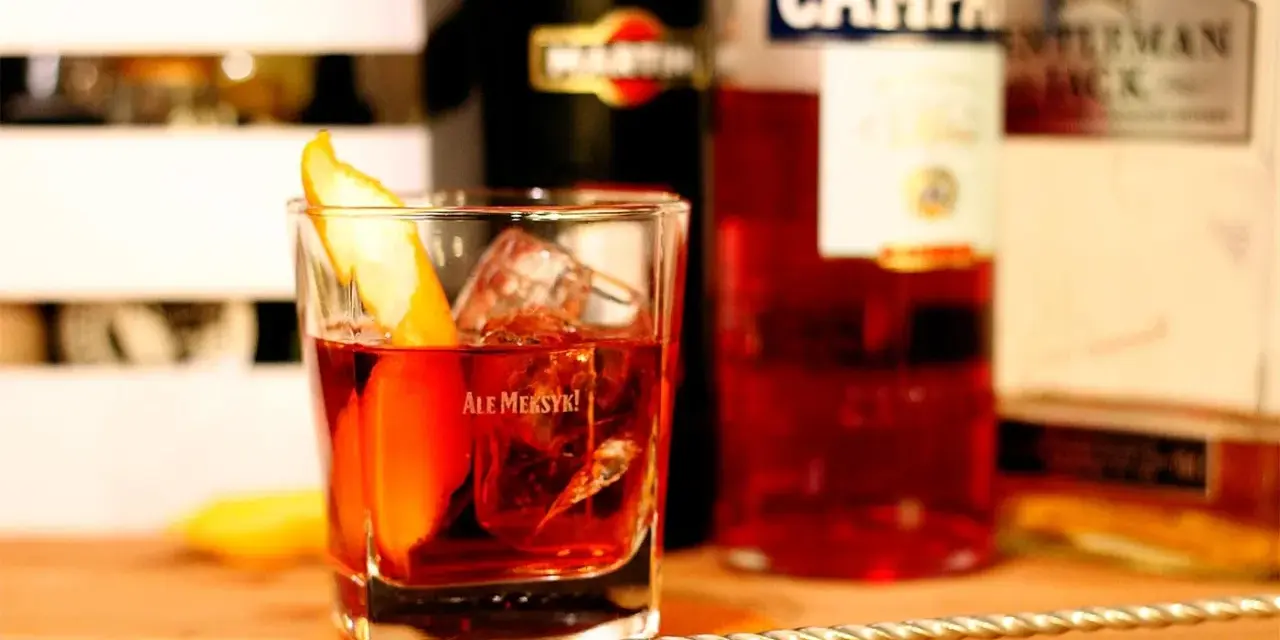 Klasyczny drink z pomarańczą i lodem. Wybór między Negroni czy Boulevardier? Na stole widać butelki Martini, Campari i Jack Daniel's.