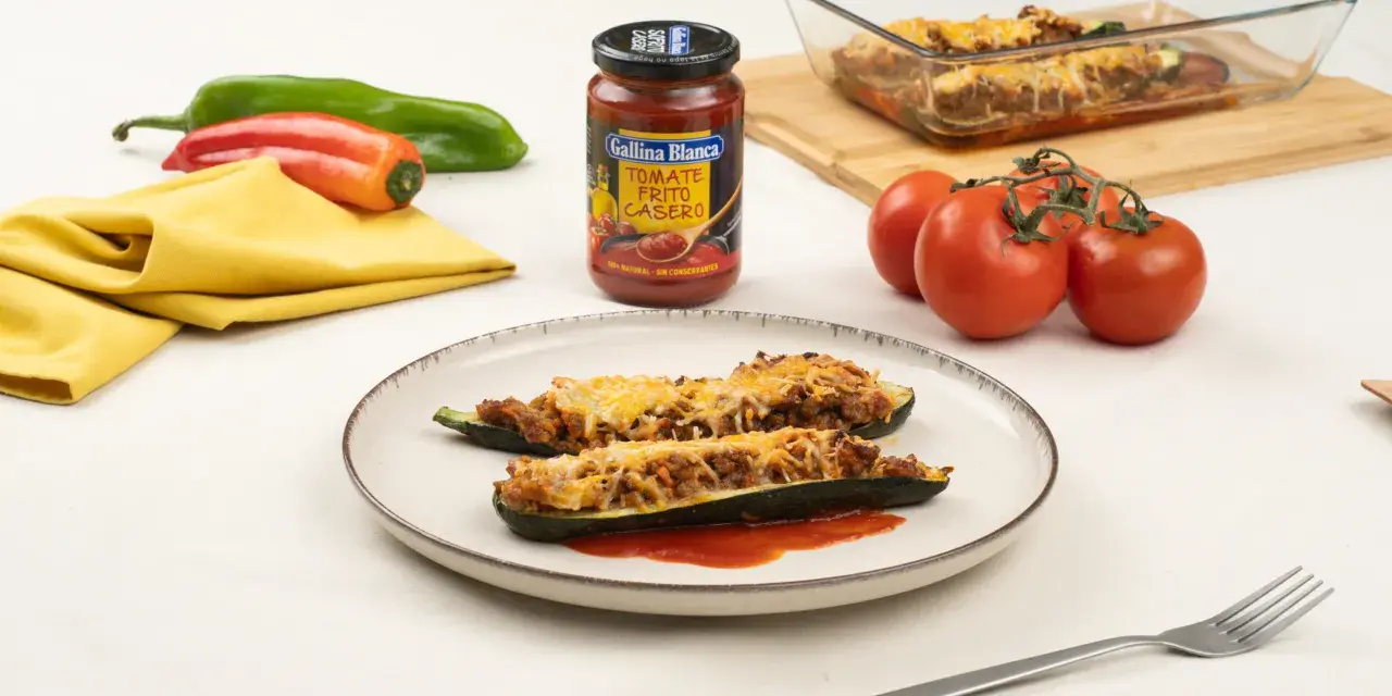 Dos calabacines rellenos de carne y gratinados con queso, servidos en un plato con tomate frito.