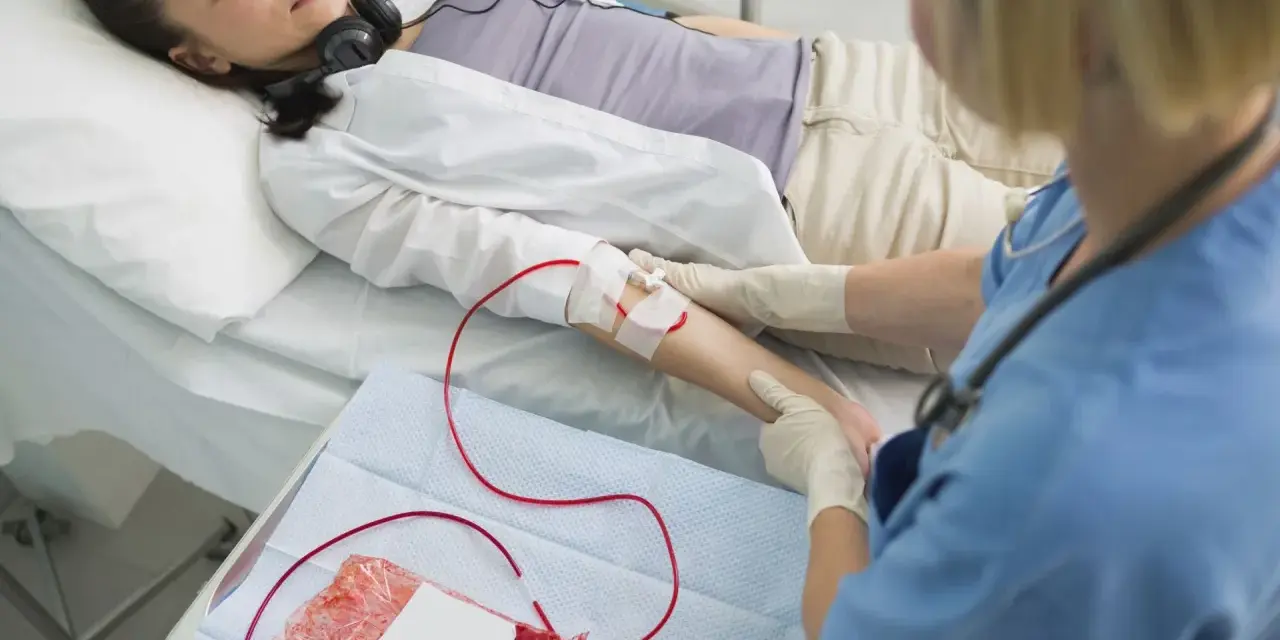 Kobieta leży w łóżku szpitalnym, słuchając muzyki przez słuchawki. Pielęgniarka wykonuje przetaczanie krwi, podłączając worek z krwią do jej ręki.
