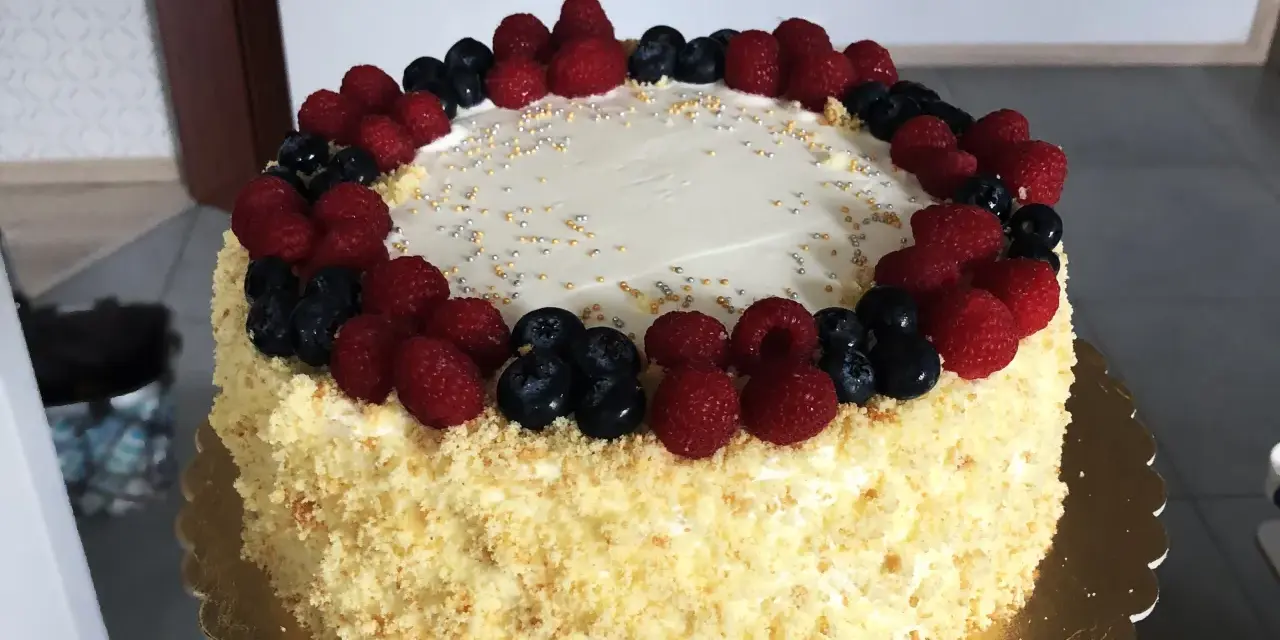 Pyszny tort śmietankowo-malinowy z mascarpone, ozdobiony świeżymi malinami i borówkami, posypany złotymi i srebrnymi kuleczkami.
