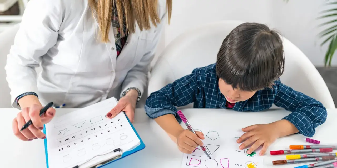 Bambino impegnato in un test per l'autismo, colora forme geometriche guidato da un'insegnante.