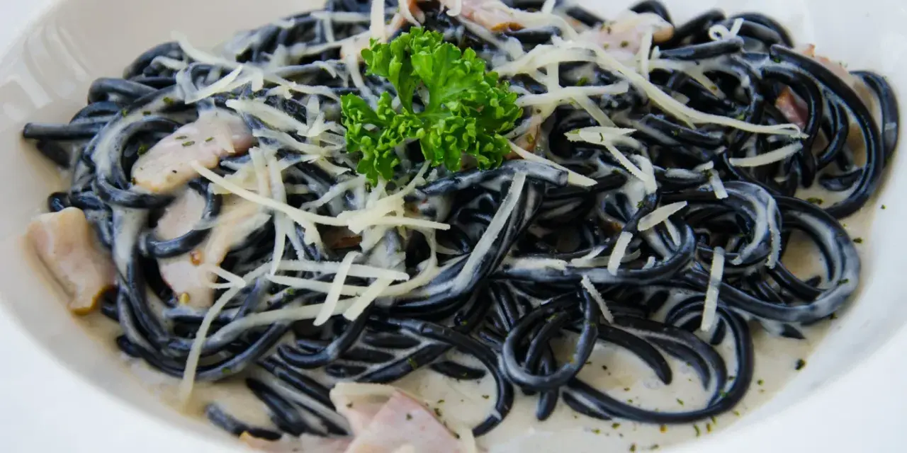 Czarny makaron spaghetti w kremowym sosie, posypany startym parmezanem i udekorowany gałązką natki pietruszki.
