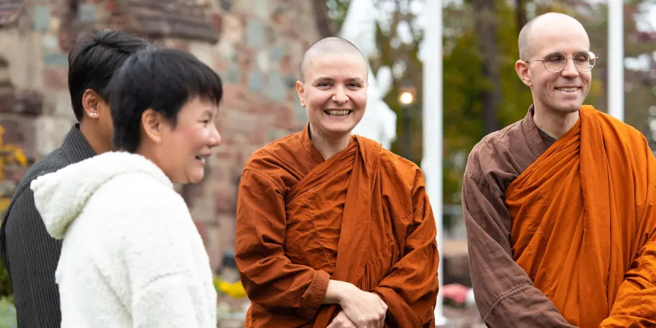 A smiling Buddhist monk in orange robes stands with others outside a church.