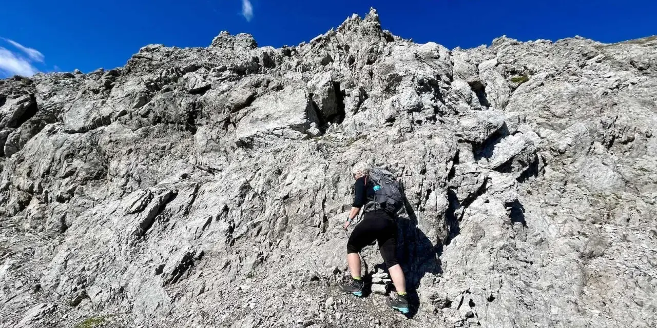 Bergsteigerin klettert über felsiges Gelände, das der UIAA-Skala entspricht. Blauer Himmel und schroffe Felsen.