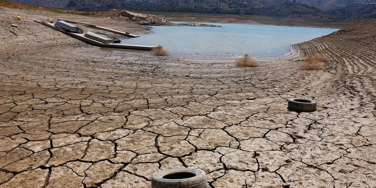 Seca tierra agrietada con un lago bajo y barcos varados. Neumáticos abandonados sugieren una contaminación natural y humana.