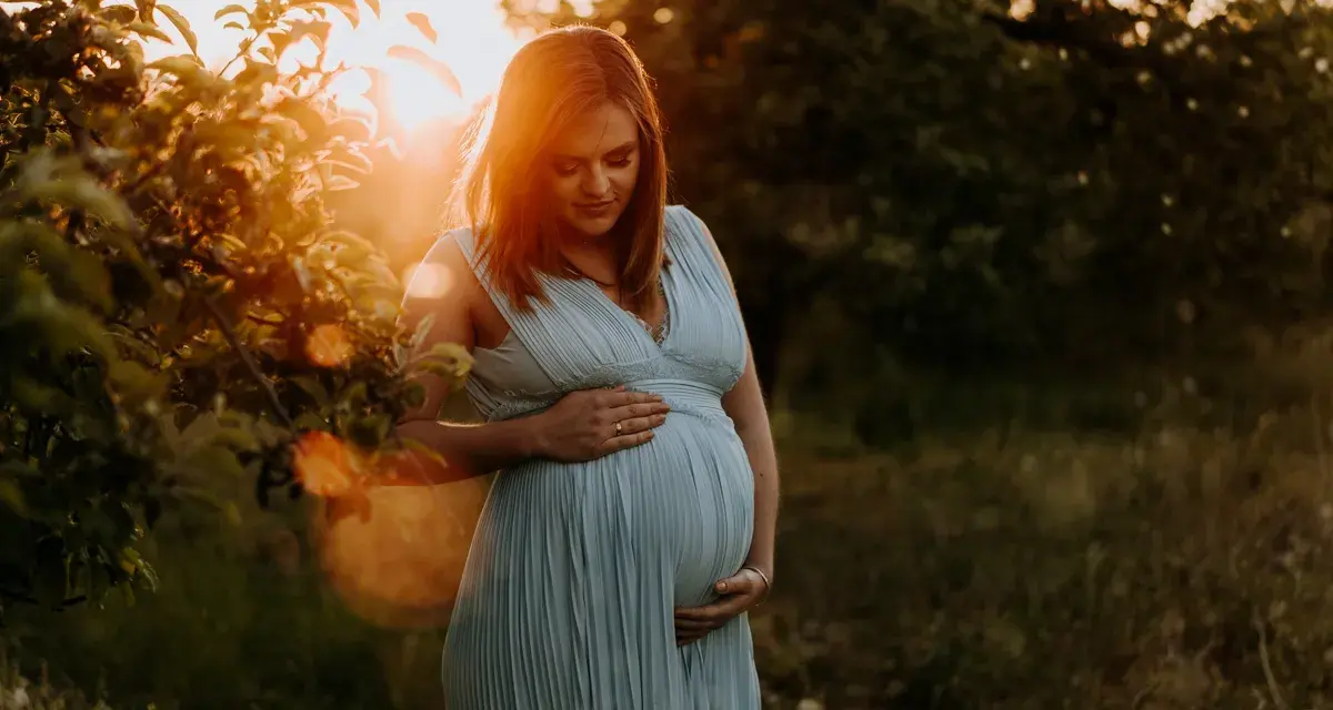 Piękne zdjęcia w ciąży. Kobieta w błękitnej sukience trzyma dłonie na brzuchu, otoczona ciepłym światłem zachodzącego słońca.