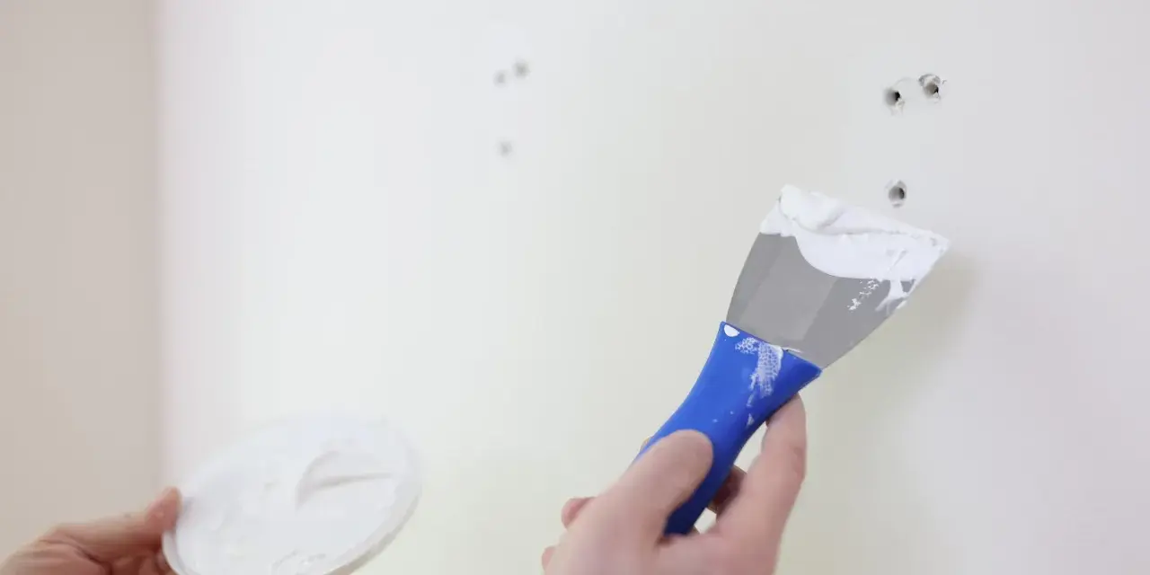 Persona aplica masilla en una pared blanca con una espátula azul para reparar agujeros.