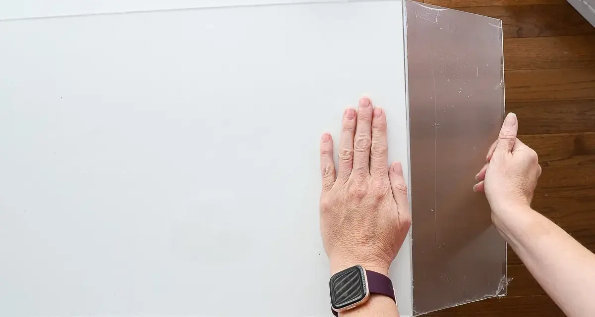Hands guide a scoring tool along a white acrylic sheet, demonstrating how to cut acrylic sheet for a project.