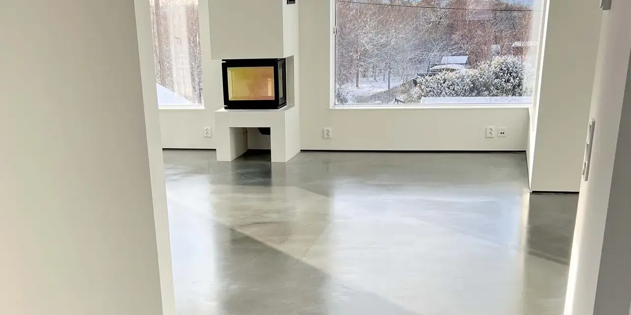 Ett modernt vardagsrum med en öppen spis och stora fönster som vetter mot en snötäckt trädgård. Golvet är ett vackert polerat betonggolv som reflekterar ljuset.