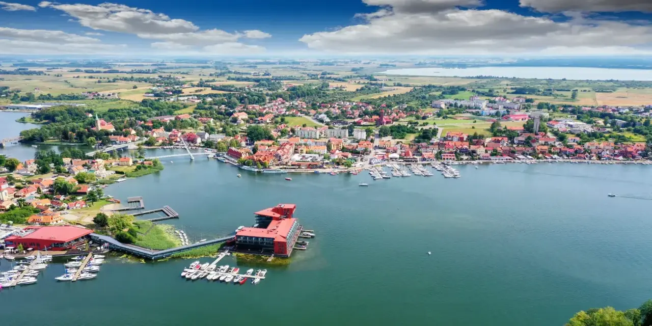 Malownicze miasta na Mazurach, które warto odwiedzić, rozciągają się wzdłuż jeziora. Widok na port z żaglówkami i zabytkową zabudowę.