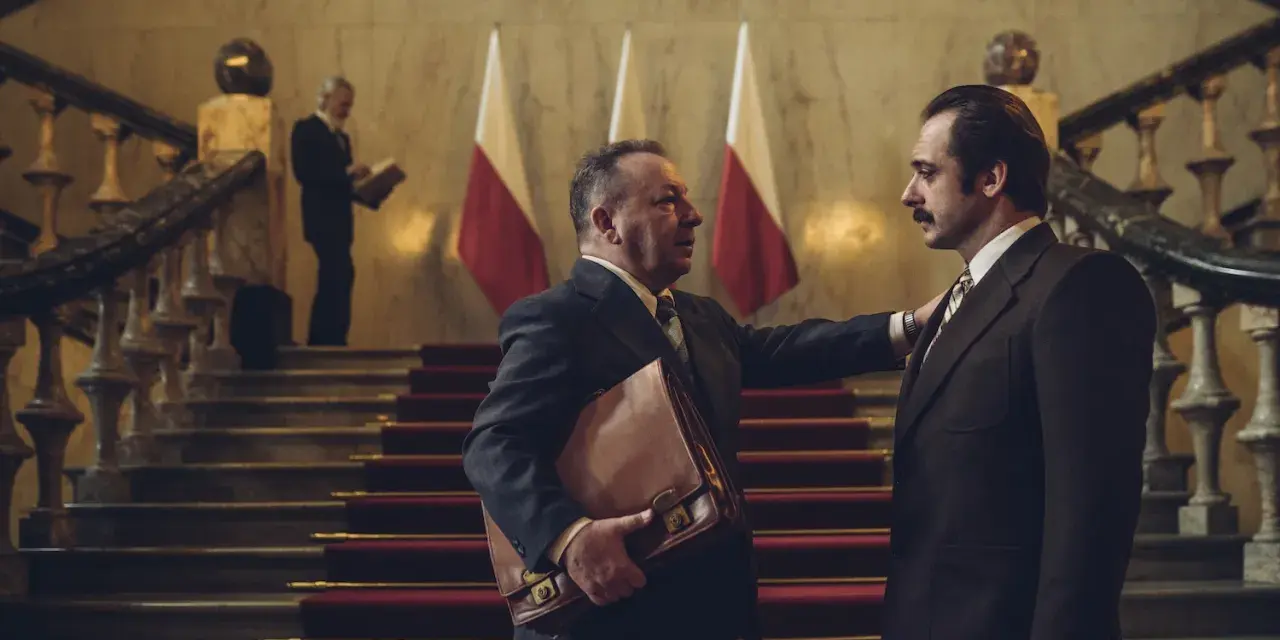 Hubert Niedziela, w garniturze, trzyma teczkę, rozmawiając z mężczyzną na schodach przed polskimi flagami.