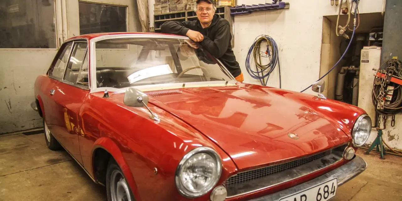 Man lutar sig mot en röd klassisk bil i garage. Lite rost syns på bilens skärmkanter.