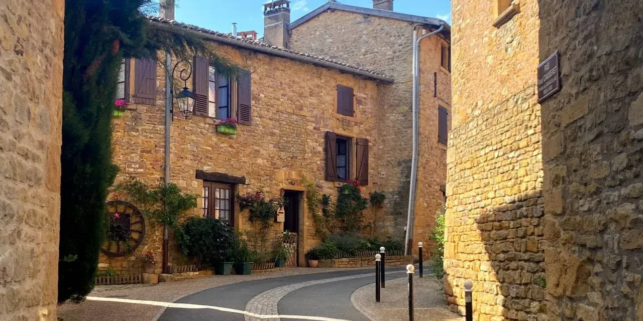 Village pittoresque aux maisons de pierre dorée, avec une ruelle sinueuse bordée de végétation luxuriante.