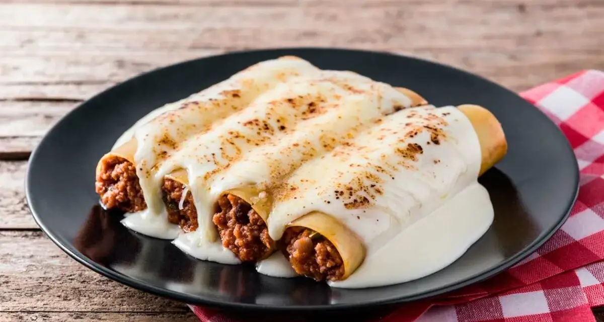 Canelones de carne picada cubiertos de queso fundido, servidos en un plato negro sobre una mesa de madera rústica.