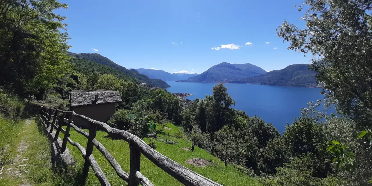 Sentiero del Viandante tappe: un casolare in pietra con tetto in ardesia e un recinto in legno rustico si affacciano su un lago blu intenso circondato da montagne verdi.