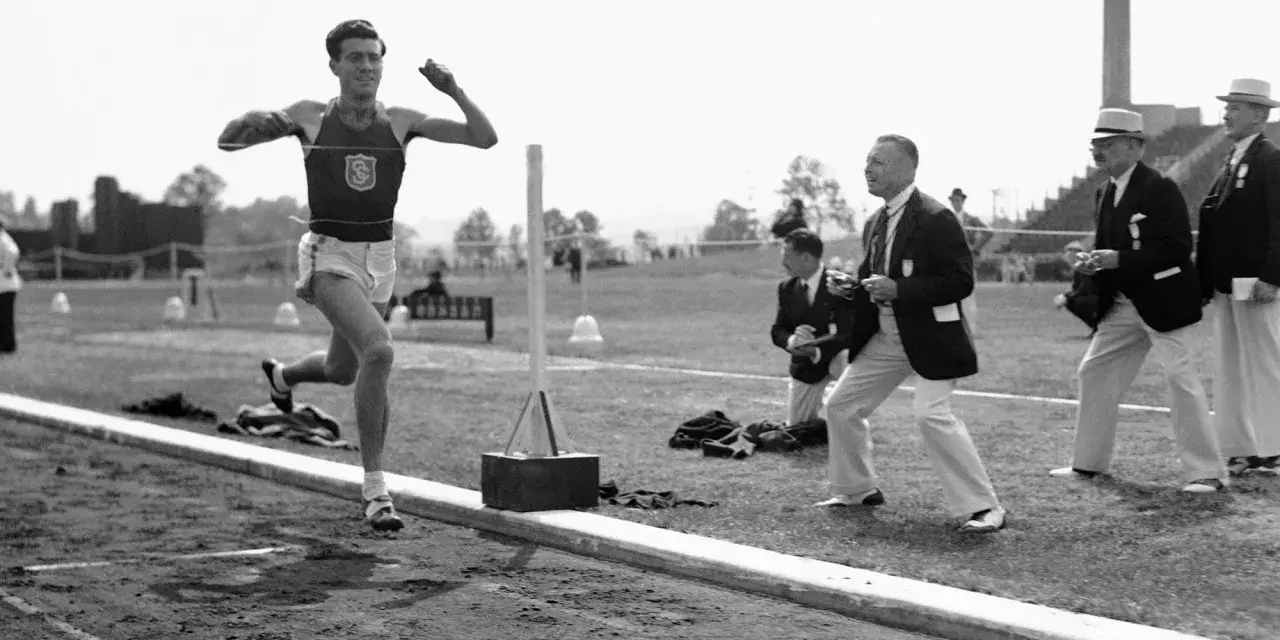 Louis Zamperini biegnie na bieżni, podczas gdy sędziowie obserwują zawody.