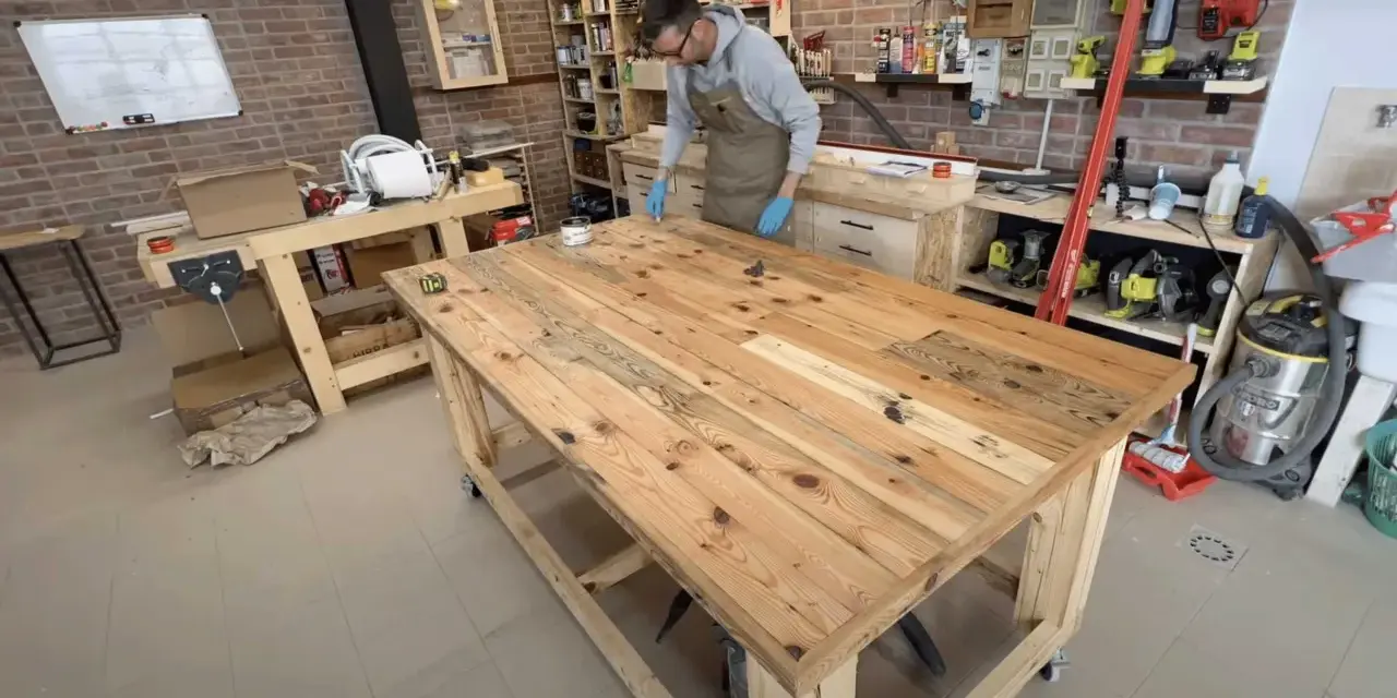 Uomo con grembiule lavora su un banco da lavoro con pallet, applicando finitura al legno. L'officina è piena di attrezzi e materiali.