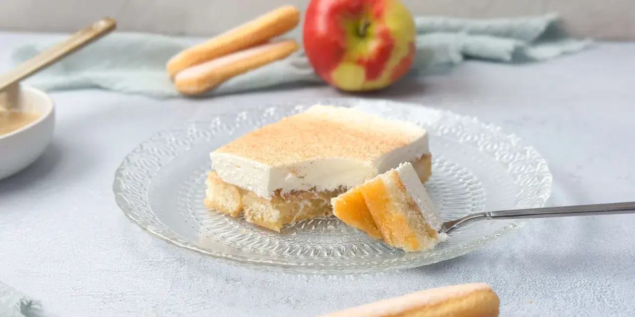 Ein Stück Apfel Schmand Kuchen mit Löffelbiskuits und einem Apfel im Hintergrund.