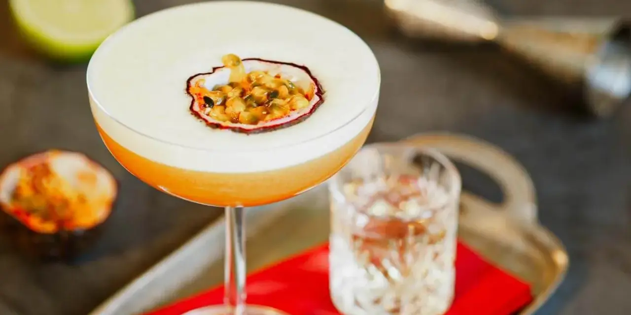 Orzeźwiający drink z martini, ozdobiony połówką marakui, obok kieliszka z wodą i połówki limonki.