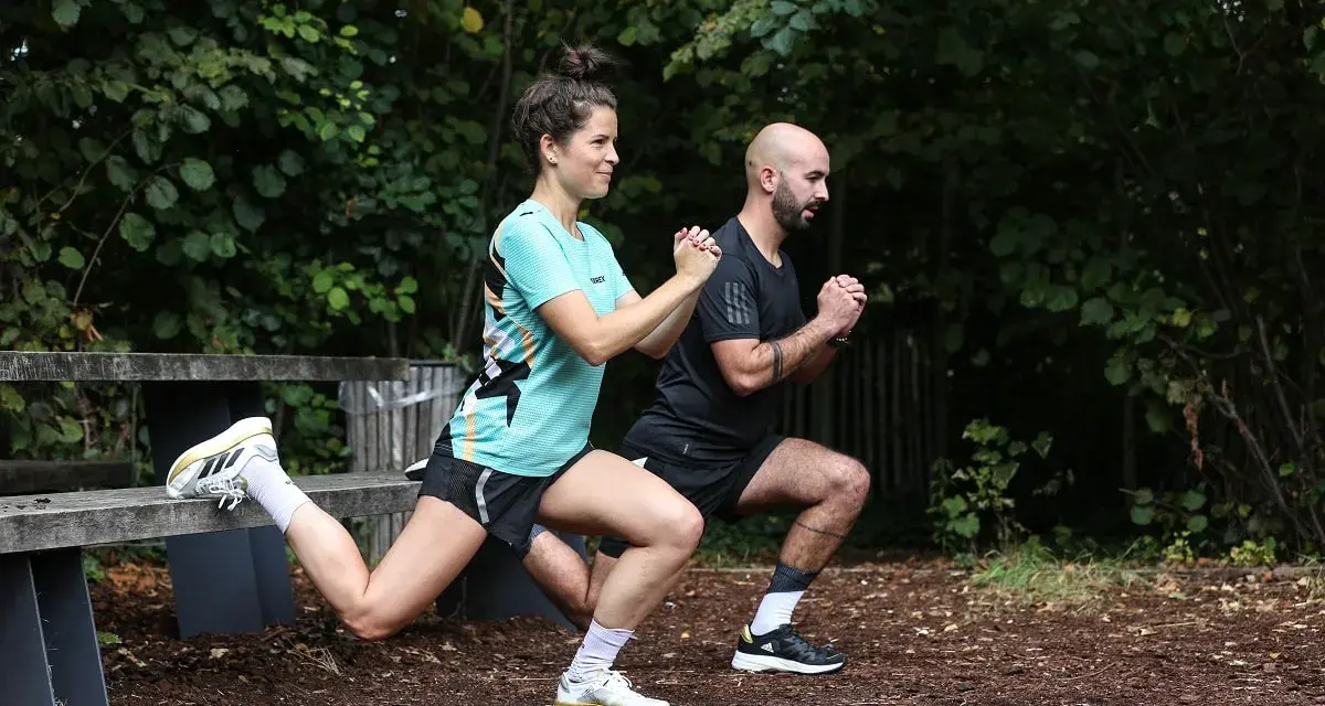 Un couple fait des exercices de renfo course à pied en extérieur, avec des fentes et des squats.