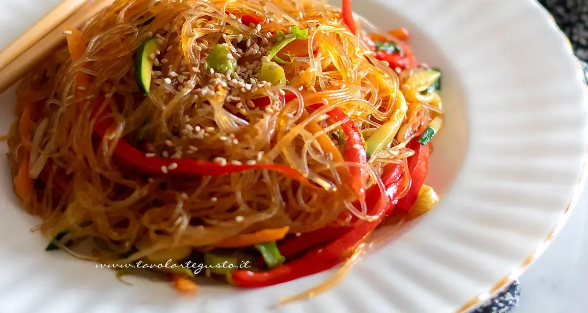 Un piatto di spaghetti di soia con verdure colorate, peperoni rossi, zucchine e carote, guarnito con semi di sesamo.