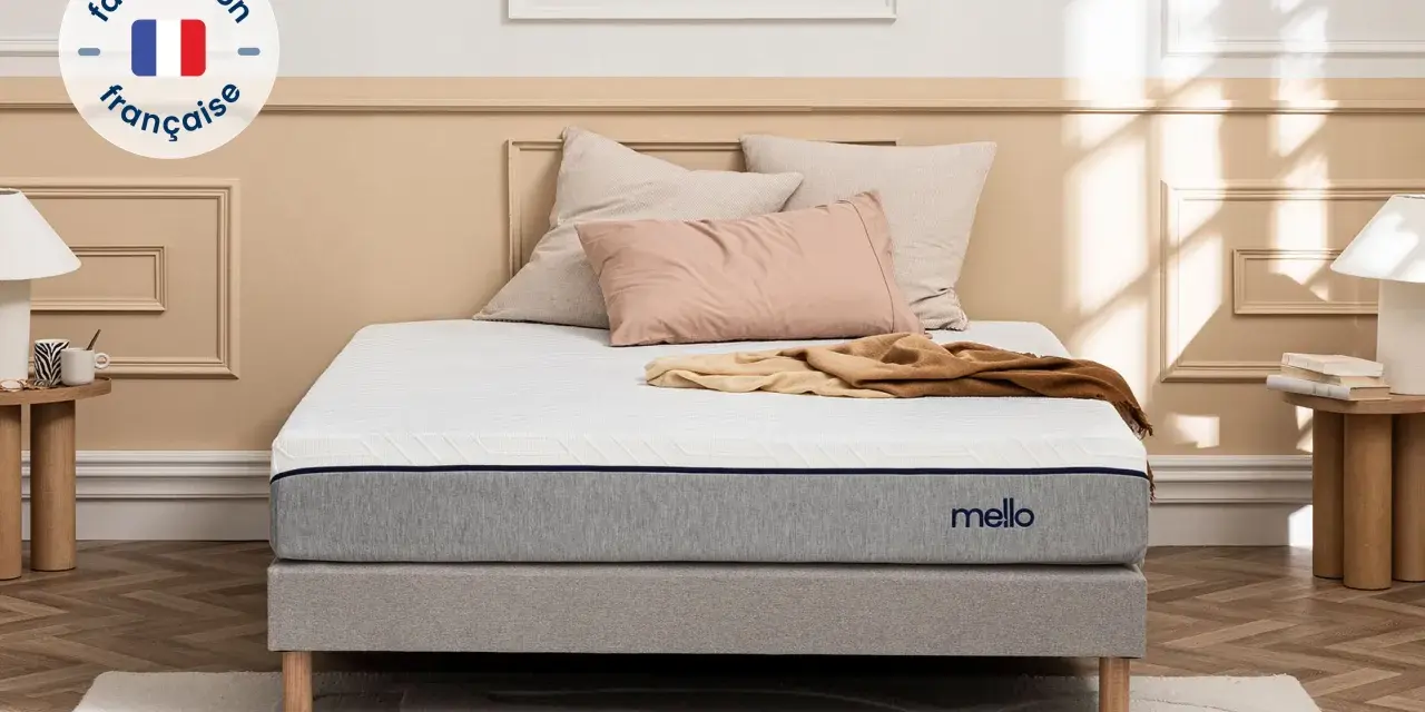 Matelas gris et blanc avec logo "mello", sur un sommier, avec des coussins et un plaid.