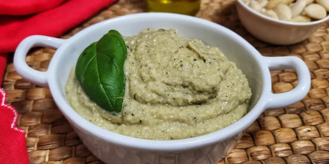 Pesto di melanzane cremoso in una ciotola bianca, guarnito con una foglia di basilico. Sullo sfondo, mandorle e olio d'oliva.