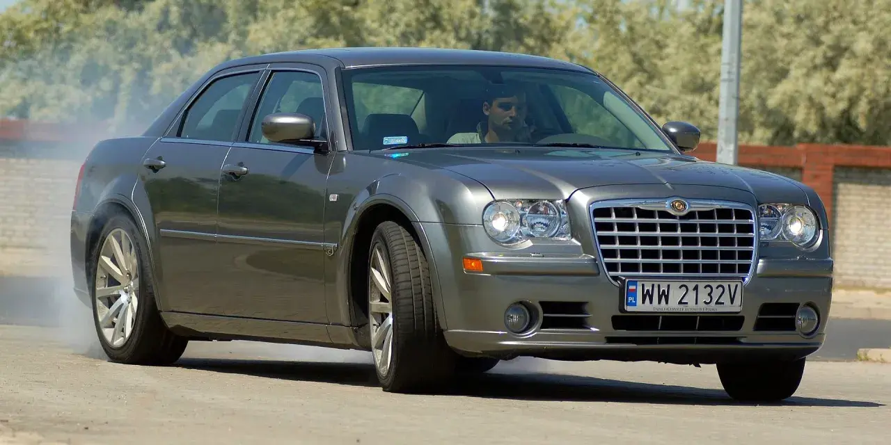 Szary Chrysler 300S z dymiącymi oponami, kierowca za kierownicą, na drodze.
