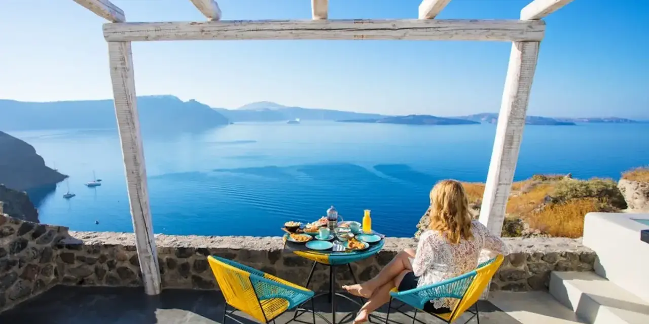 Mujer desayunando en balcón con vista al mar azul y montañas.