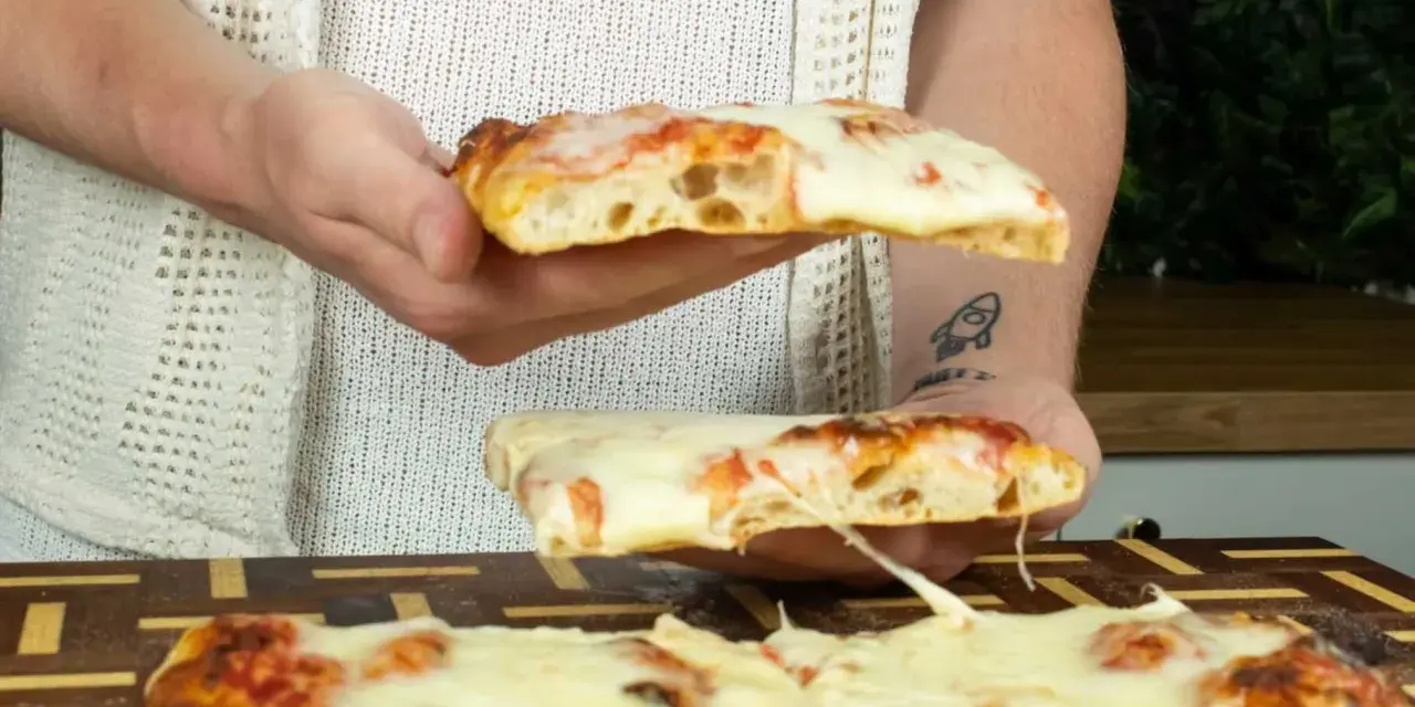 Due fette di pizza in teglia, con mozzarella filante e crosta dorata, vengono sollevate da mani.
