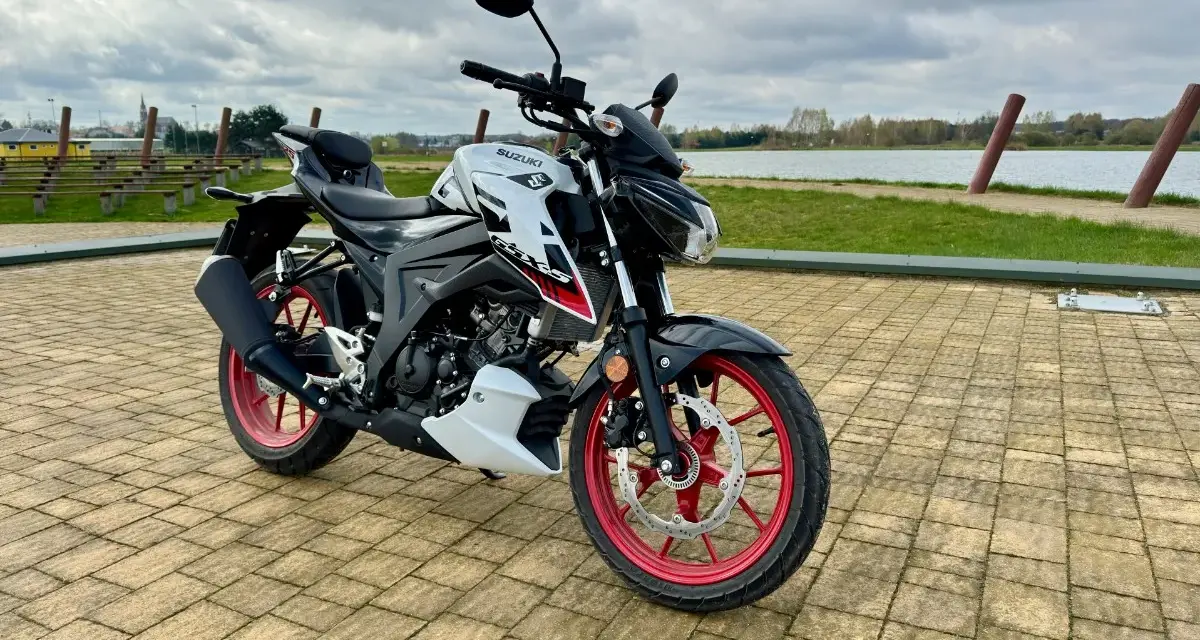 Biało-czarny Suzuki GSX S125 z czerwonymi felgami stoi na wybrukowanym placu nad jeziorem.