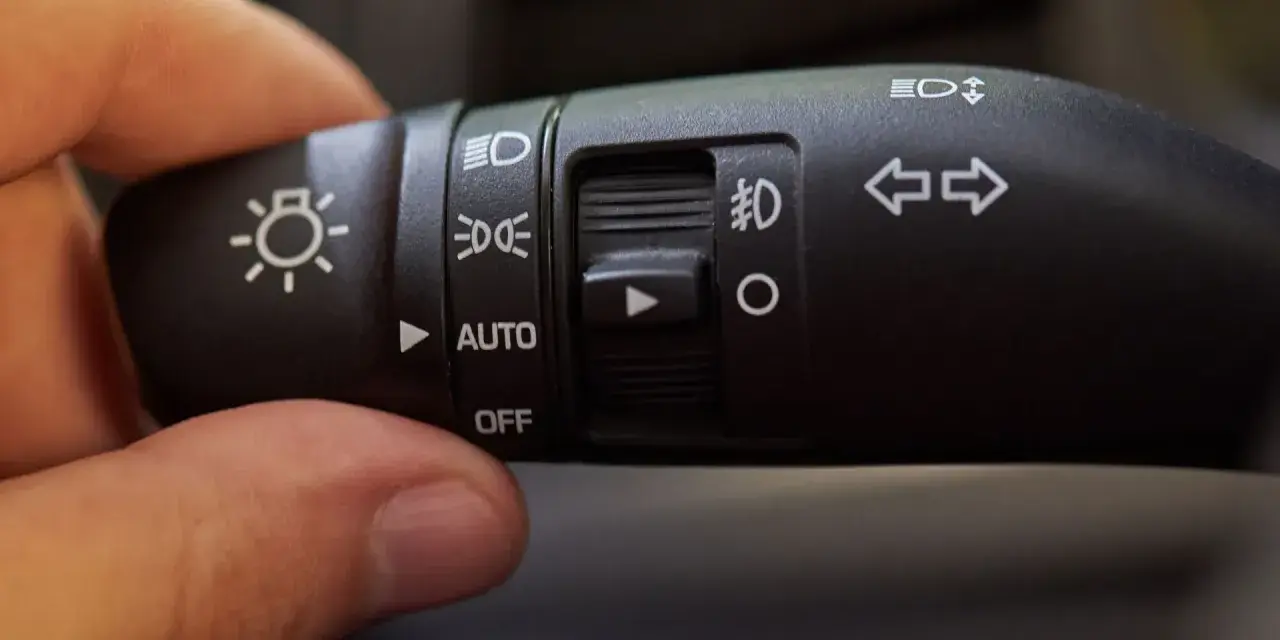 Dłoń reguluje przełącznik świateł auto. Widoczne symbole oznaczają światła drogowe, mijania, postojowe i automatyczne.