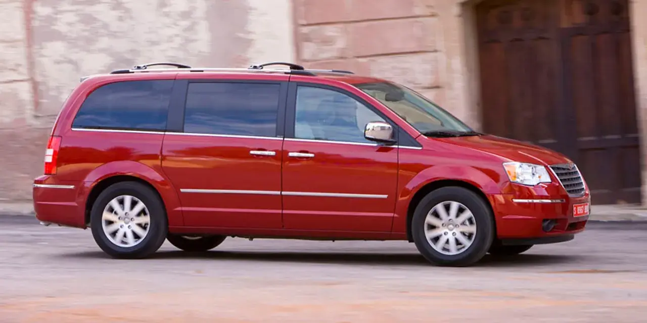 Czerwony Chrysler Grand Voyager w ruchu, z przyciemnianymi szybami i relingami dachowymi, przemierza ulicę obok kamiennej fasady.