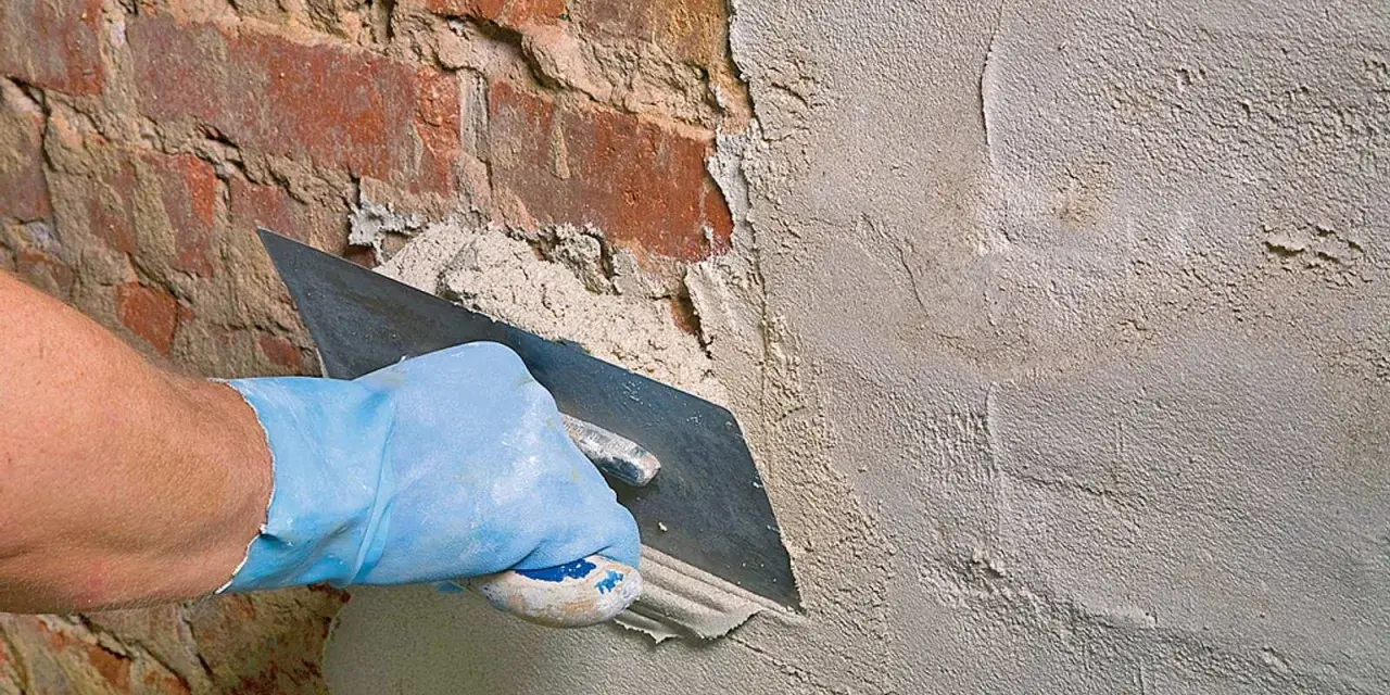 Hand mit blauer Handschuh glättet mit Kelle den **Sanierputz Keller** auf rotem Ziegelmauerwerk.