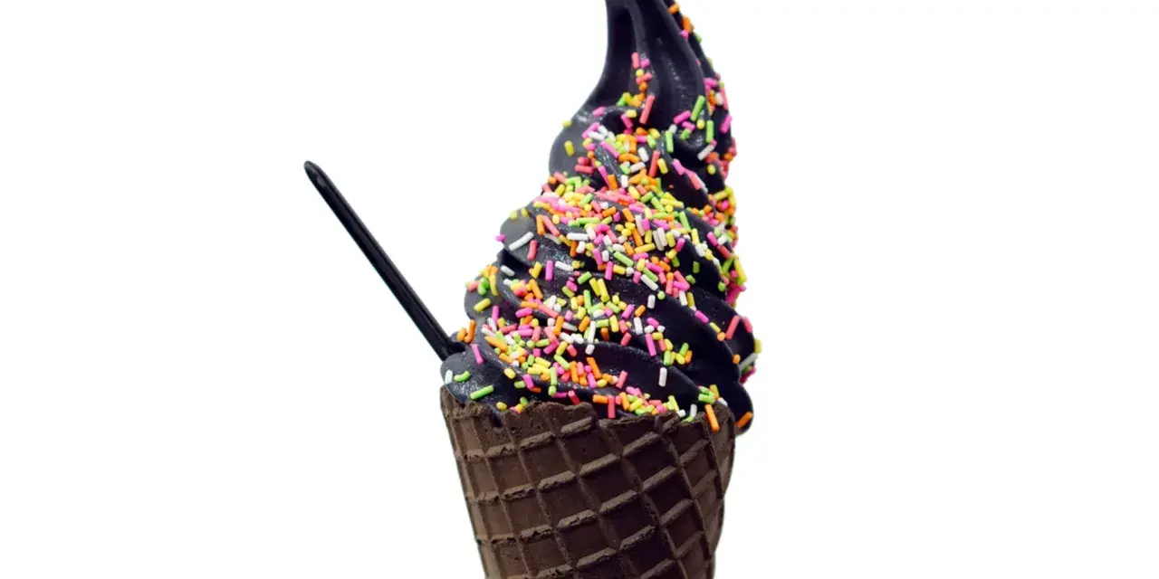 Schwarzes Eis in einer Waffel, bestreut mit bunten Streuseln. Ein schwarzer Löffel steckt darin.