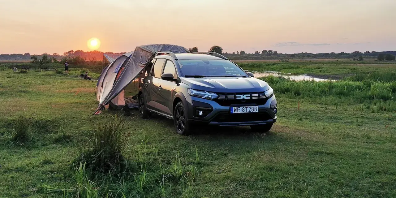 Dacia Jogger Camper rozłożony na biwaku o zachodzie słońca. Namiot połączony z autem, otoczony łąką i wodą.
