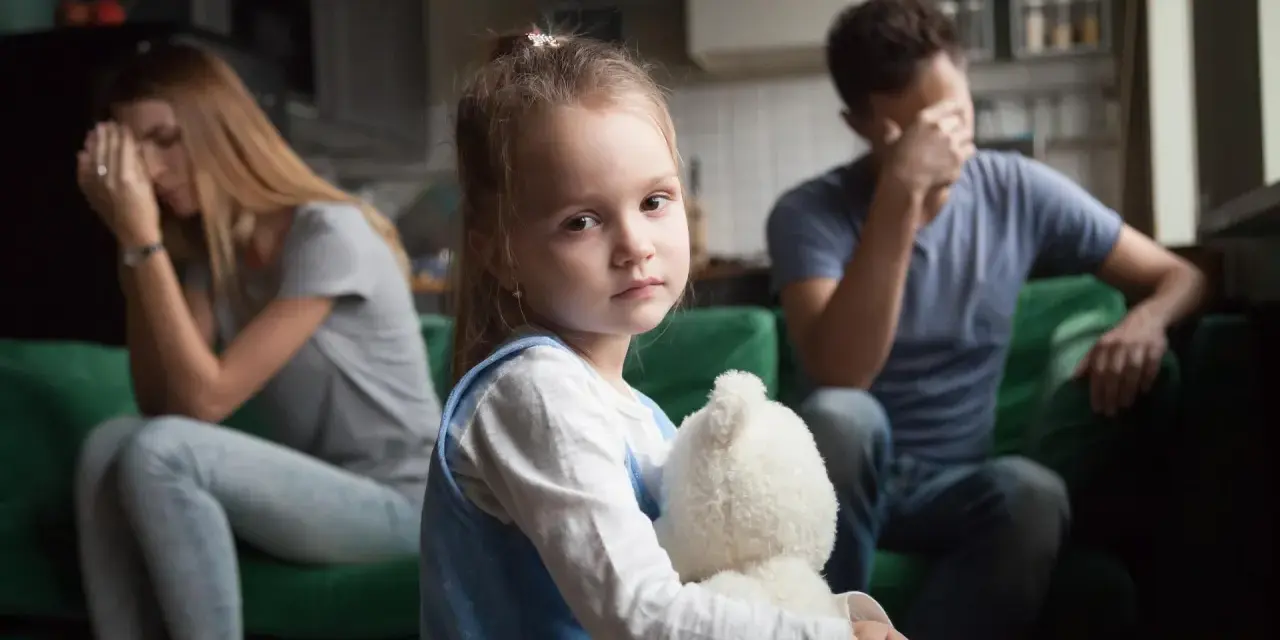 Smutna dziewczynka z misiem w objęciach, podczas gdy rodzice w tle przeżywają trudne chwile. Temat: rozwód a dzieci.
