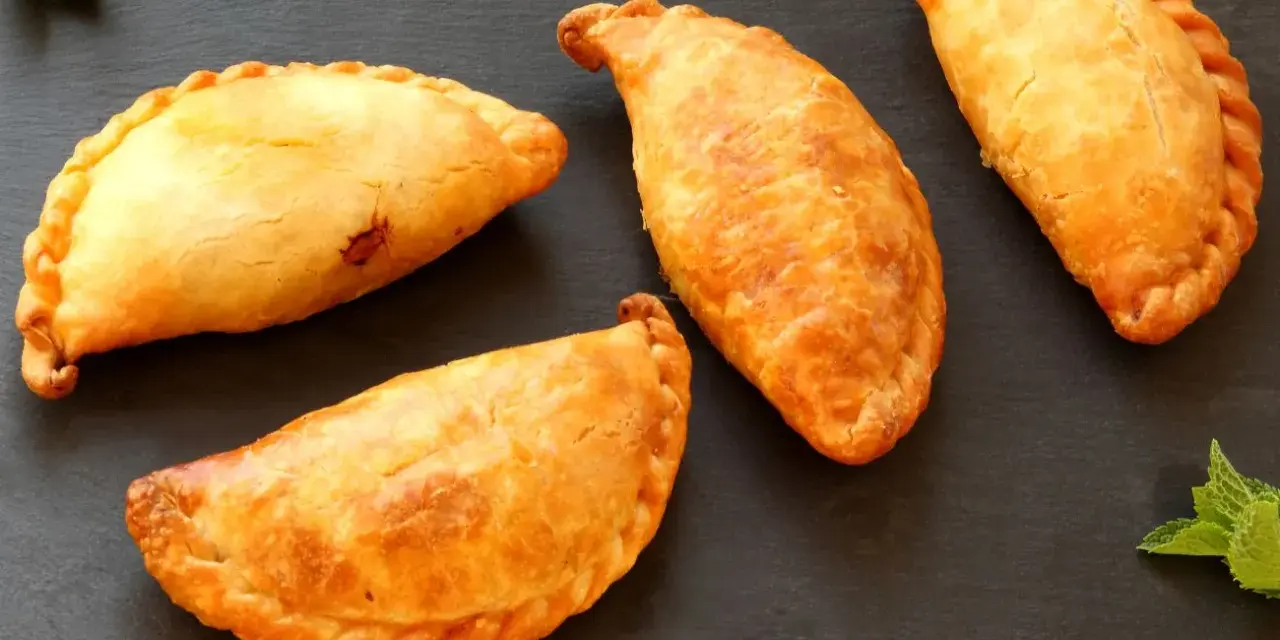 Cuatro empanadillas doradas y crujientes dispuestas sobre una superficie oscura, con hojas de menta a los lados.