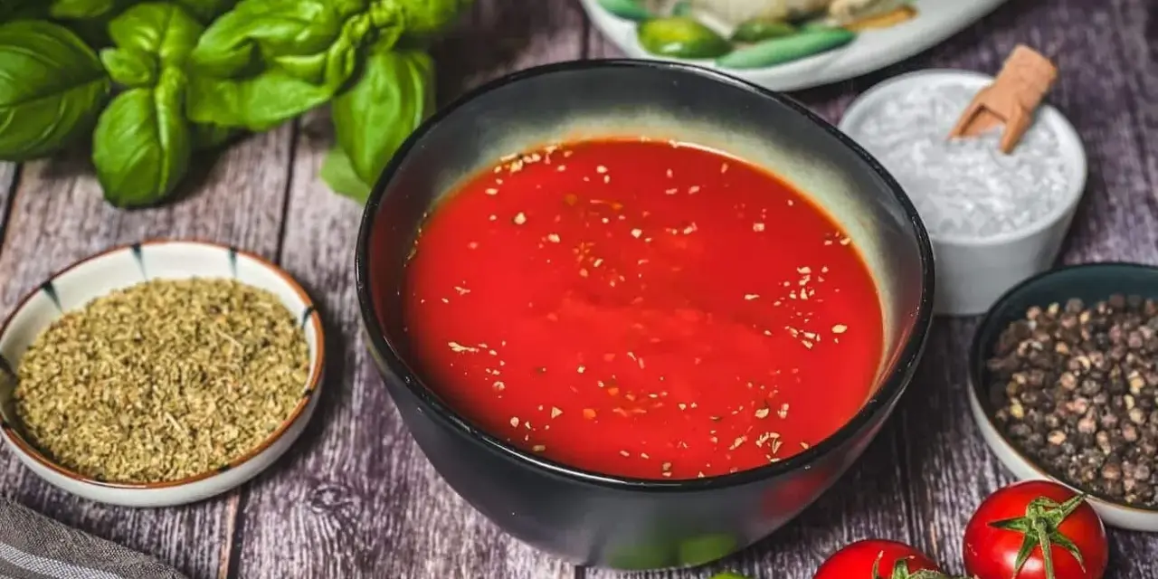 Schüssel mit leuchtend roter Pizza Sauce, umgeben von frischem Basilikum, Tomaten, Oregano, Salz und Pfeffer – wie in der Pizzeria.